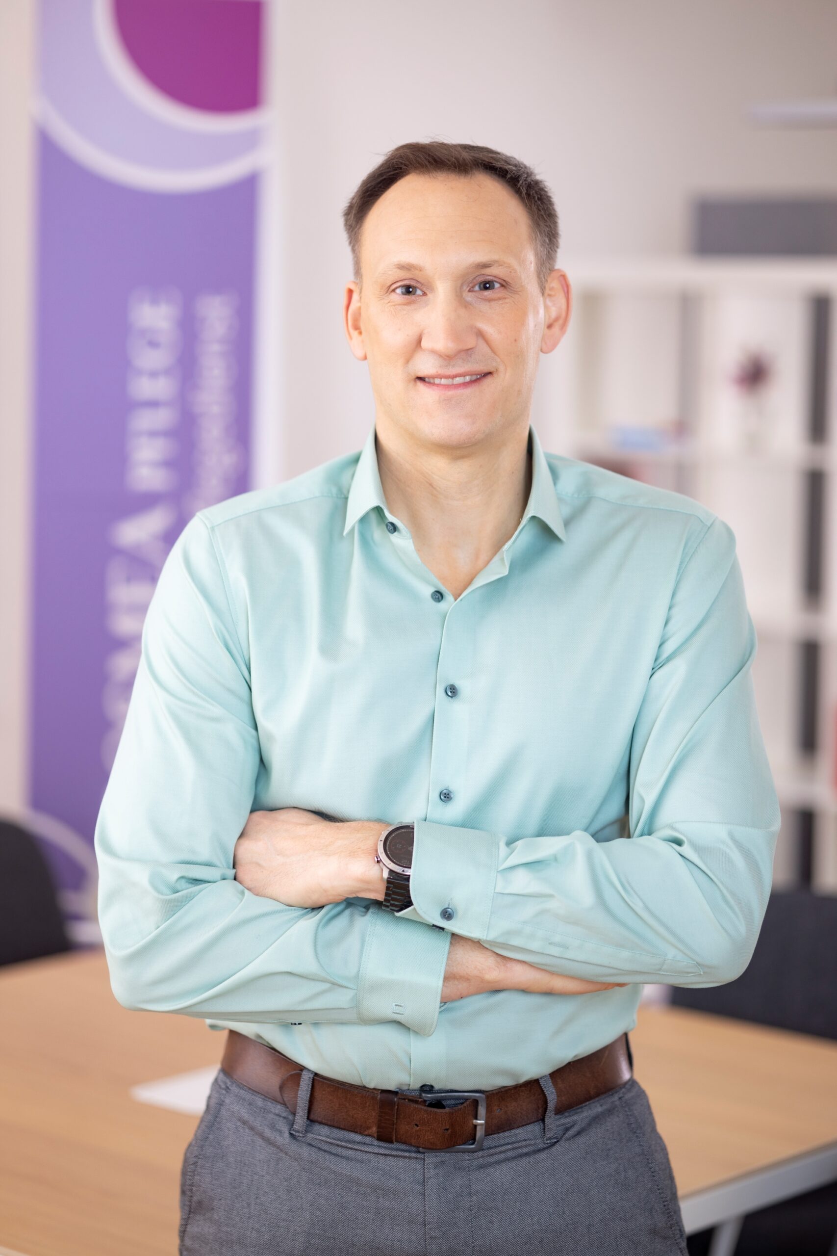 Portrait von Andreas Eichelberger, Geschäftsführer von Cosmea. Beschreibung: türkises Hemd, Arme verschränkt, freundliches Lächeln, kurze braune Haare, Cosmea Pflege Plakat im Hintergrund.