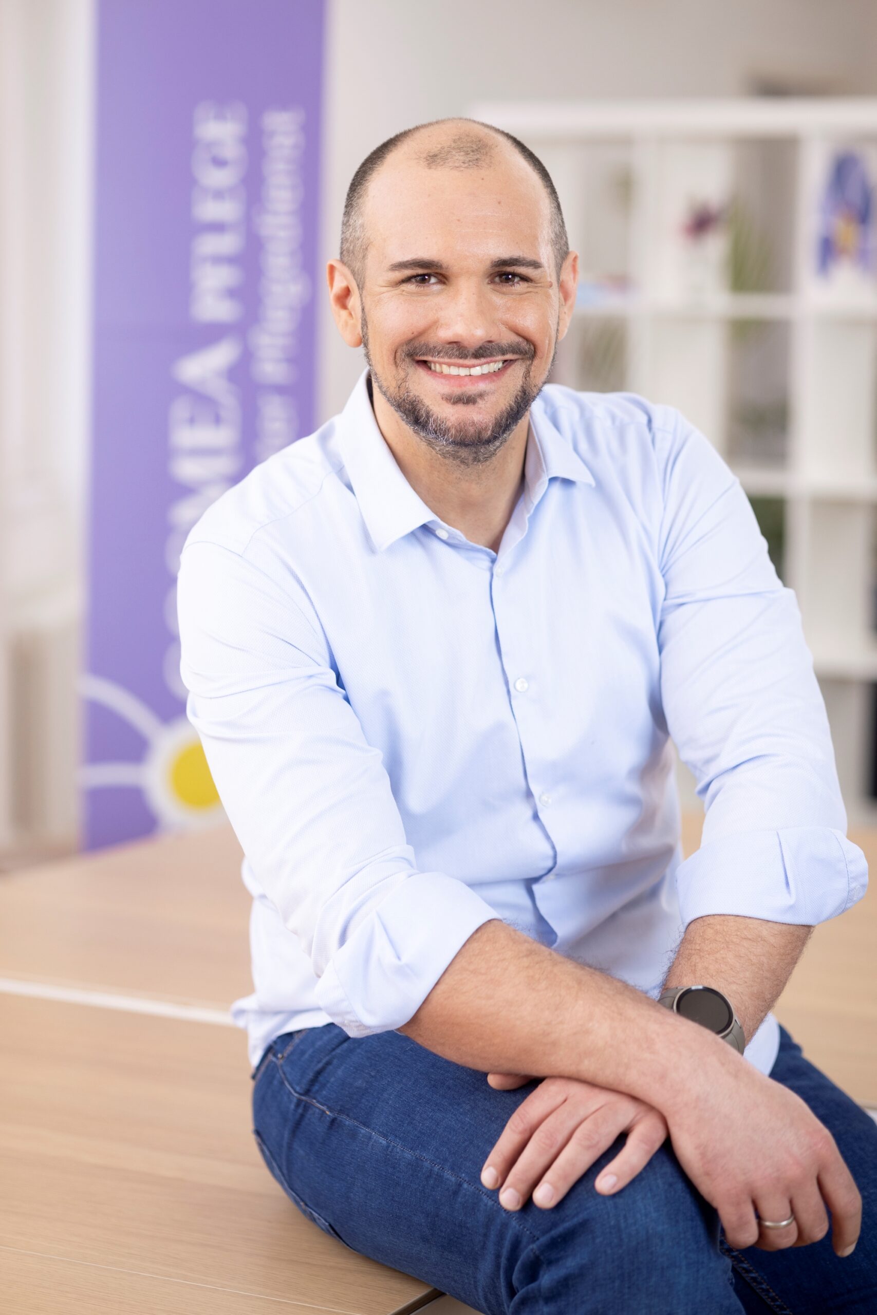 Chris Scholze, Leiter der IT Abteilung von Cosmea Pflege. Beschreibung: locker sitzend am Tisch, Hände übereinander liegend, dunkelbrauner Bart und Haare, breites Lächeln.