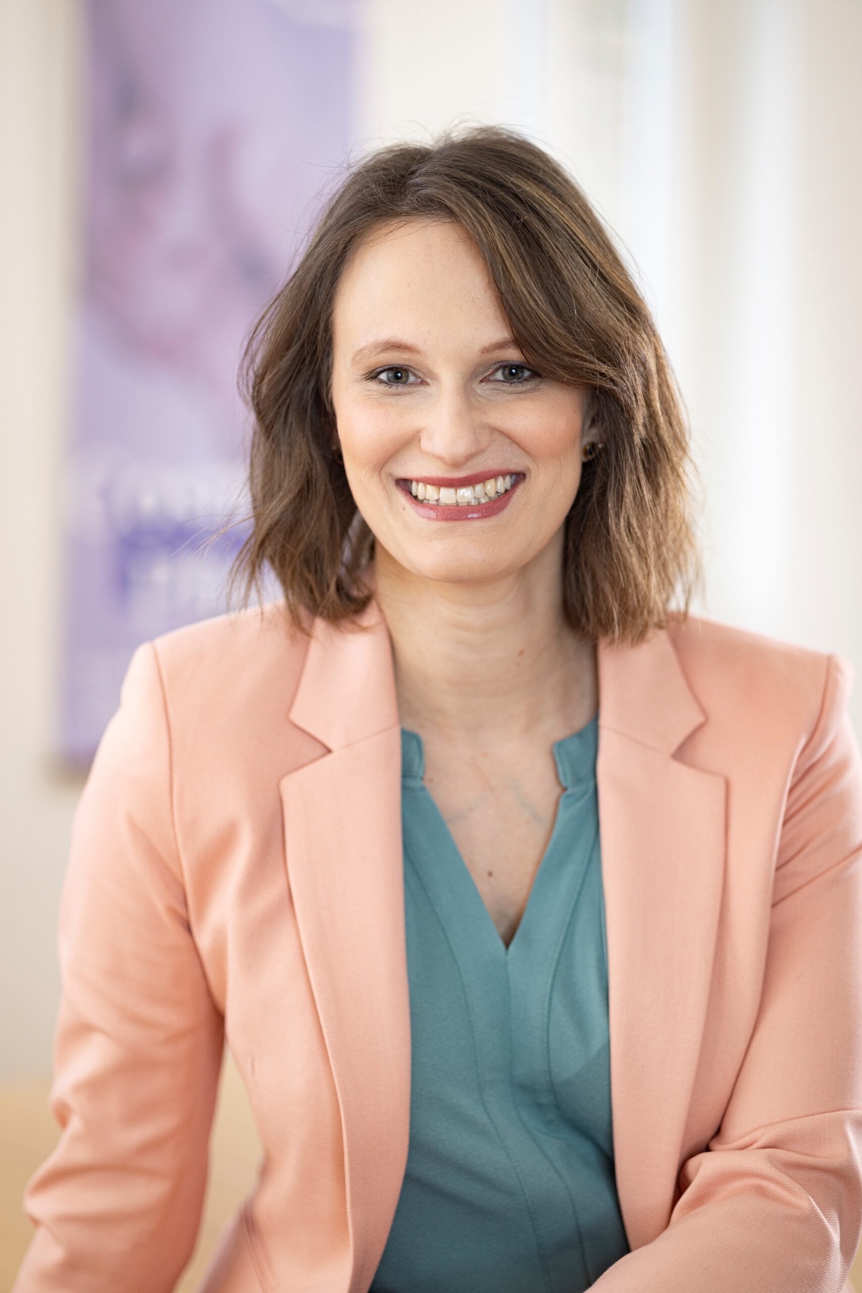 Portrait von Sandra Gehm, Leiterin der Finanzabteilung. Beschreibung: Hellrosa Sakko über türkise Bluse, Schulterlange hellbraune Haare, freundliches breites Lächeln, Cosmea Pflege Banner im Hintergrund