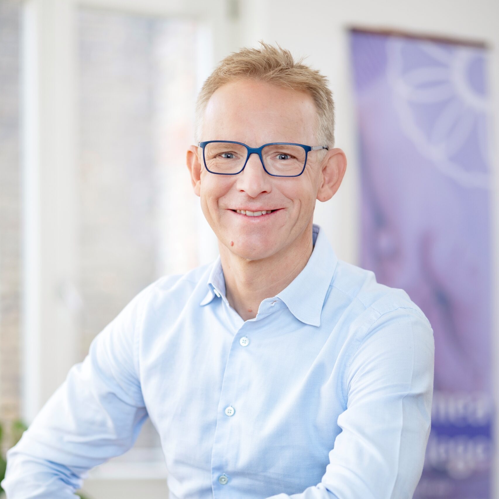 Portrait von Tomas Aubell, Geschäftsführer von Cosmea Pflege. Beschreibung: Blonde Haare, Brille, hellblaues Hemd, freundliches Lächeln